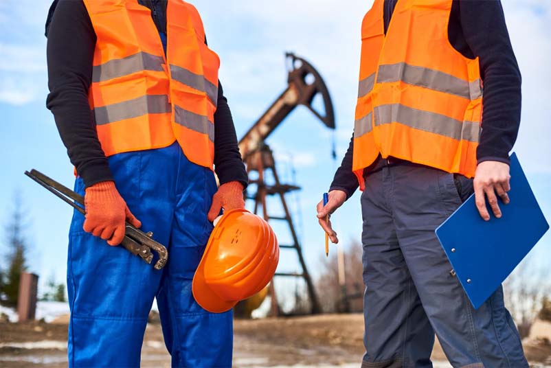 Two construction workers are looking at each other. One has a blue clipboard, and the other is holding a hard hat and a piece of metal