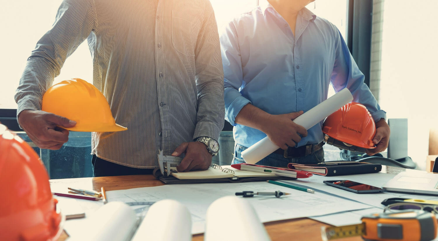 Two business professionals are holding yellow and orange hard hats, plans, and drawing tools facing a table, filled with more plans and drawing tools